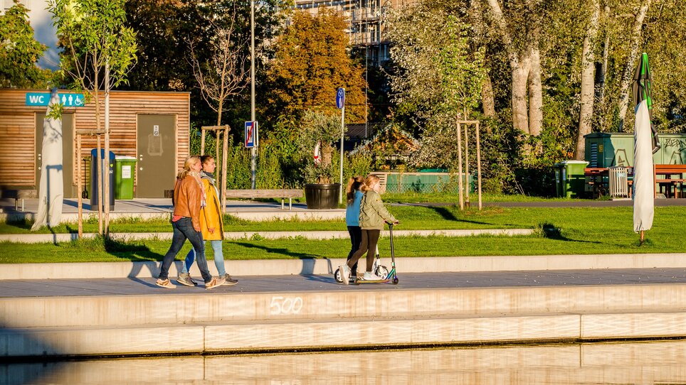 Zwei Frauen spazieren am Ufer der Mur mit zwei Mädchen auf Rollern. | © Graz Tourismus