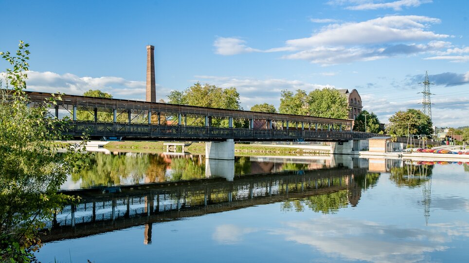 Eine Brücke über die ruhige Mur, umgeben von Bäumen und historischen Gebäuden. | © Graz Tourismus