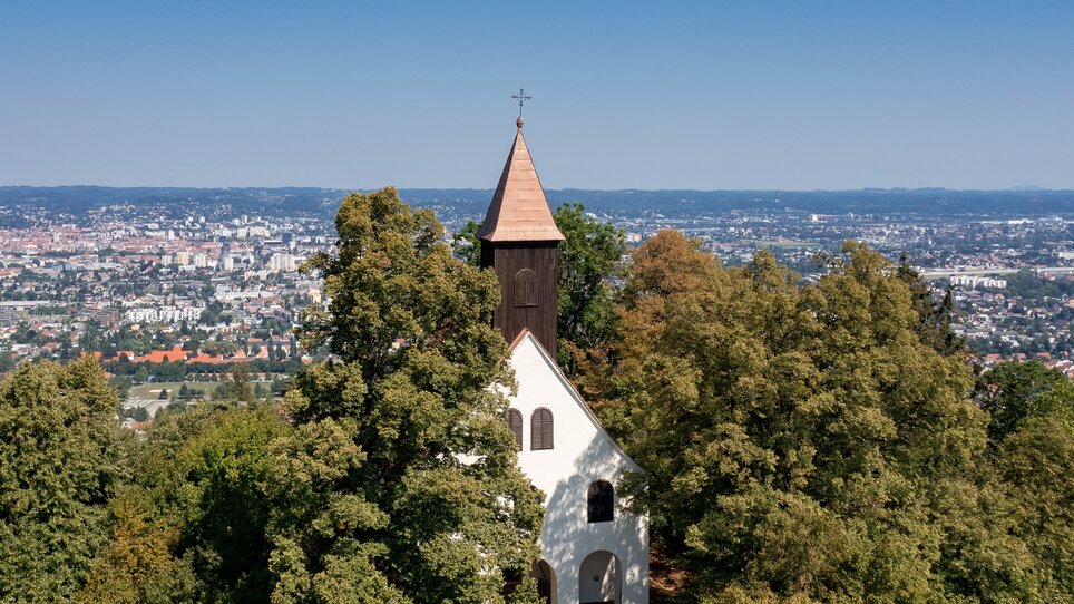The church of St John and Paul - surrounded by trees with a view of Graz. | © Graz Tourismus