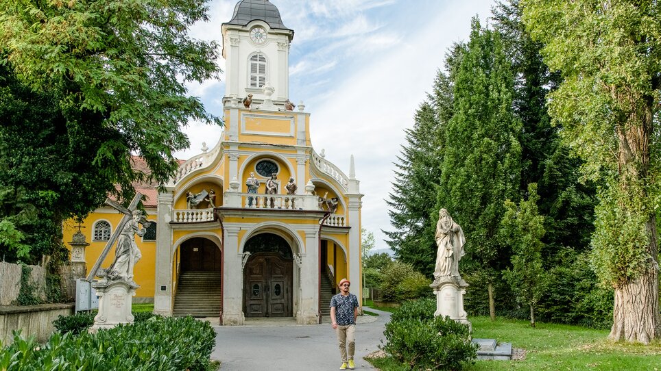 Ein Mann steht vor der Kalvarienbergkirche - einer gelben Kirche mit Statuen davor und Bäumen.