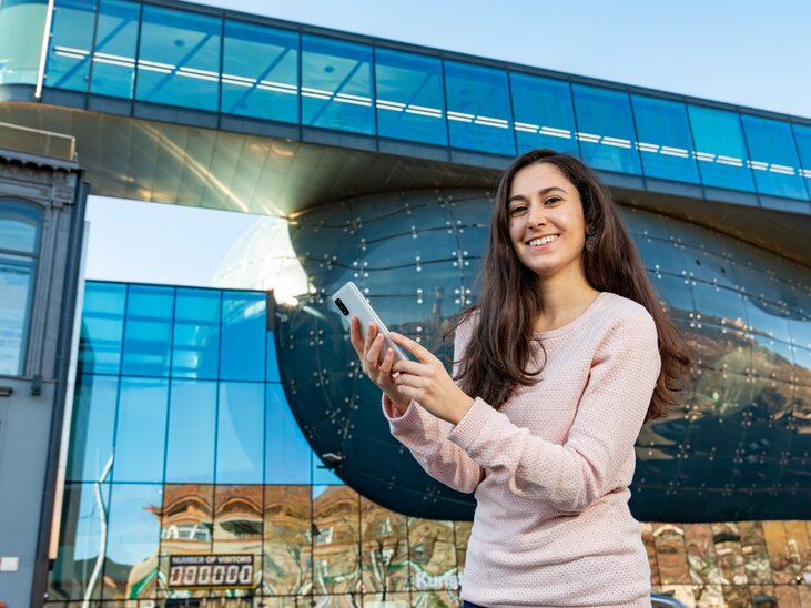 Eine junge Frau lächelt und hält ihr Smartphone vor einem reflektierenden Gebäude in Graz. | © Graz Tourismus - Harry Schiffer