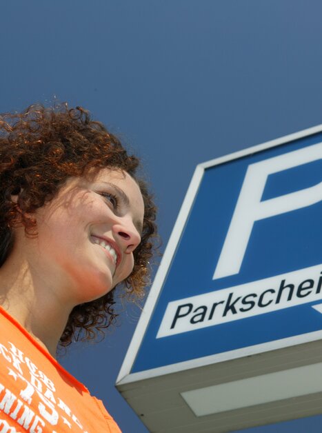 Eine Frau lächelt neben einem Schild mit dem Wort 'Parkschein', blauer Himmel im Hintergrund. | © Graz Tourismus - Harry Schiffer