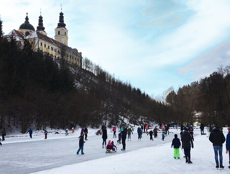 People enjoy ice skating in front of a monastery in a winter landscape. | © Stadt Graz - Sportamt