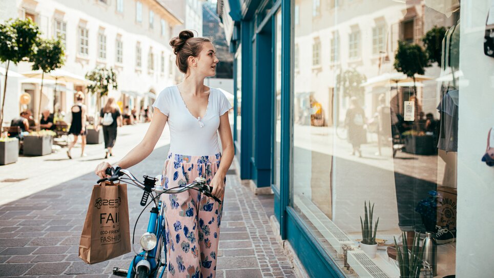 Eine junge Frau mit Blumenhose steht mit ihrem Fahrrad vor einem Schaufenster in Graz. | © Graz Tourismus