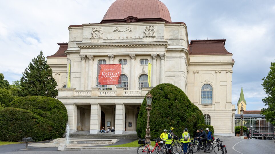 Das Opernhaus in Graz mit Radfahrern davor und einem großen Plakat. | © Graz Tourismus - Harry Schiffer