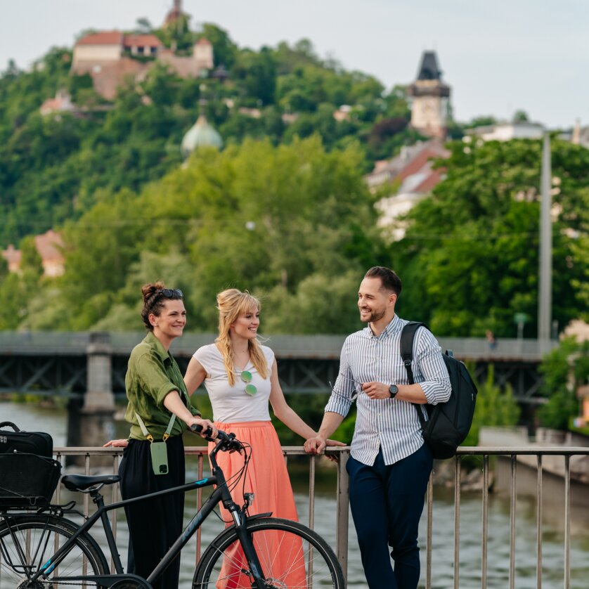Three people with bicycles in front of the Mur and the Graz Clock Tower. | © Graz Tourismus - Mias Photoart
