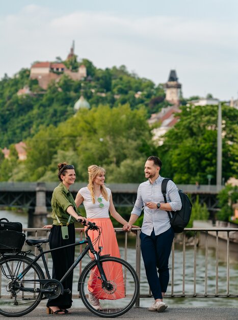 Three people with bicycles in front of the Mur and the Graz Clock Tower. | © Graz Tourismus - Mias Photoart