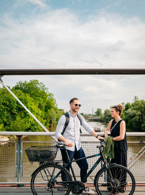 Ein Paar unterhält sich auf einer Brücke über der Mur. Vor dem Mann steht ein Fahrrad. | © Graz Tourismus - Mias Photoart