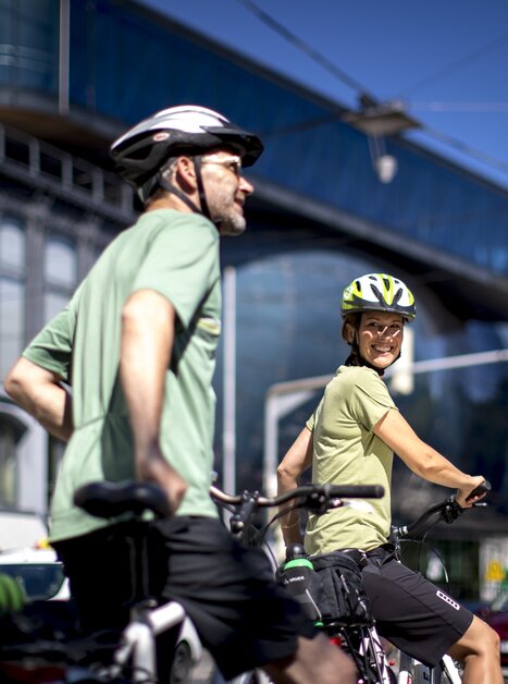 Due ciclisti sorridenti a Graz. Architettura moderna sullo sfondo. | © Graz Tourismus - Tom Lamm