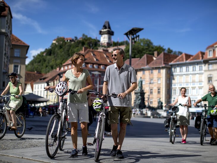 Coppia e altri ciclisti a Graz, con la Torre dell'Orologio di Graz sullo sfondo. | © Graz Tourismus - Tom Lamm