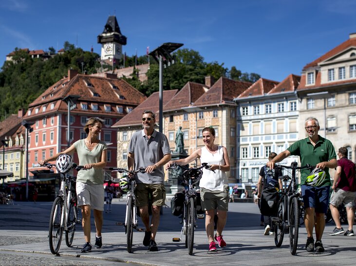 Four cyclists in Graz with the Graz Clock Tower in the background. | © Graz Tourismus - Tom Lamm