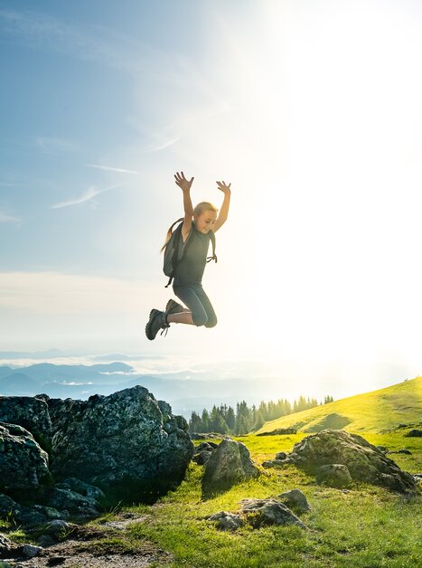 Ein Mädchen springt fröhlich in die Luft vor einer sonnigen Berglandschaft. | © Region Graz - Mias Photoart-43