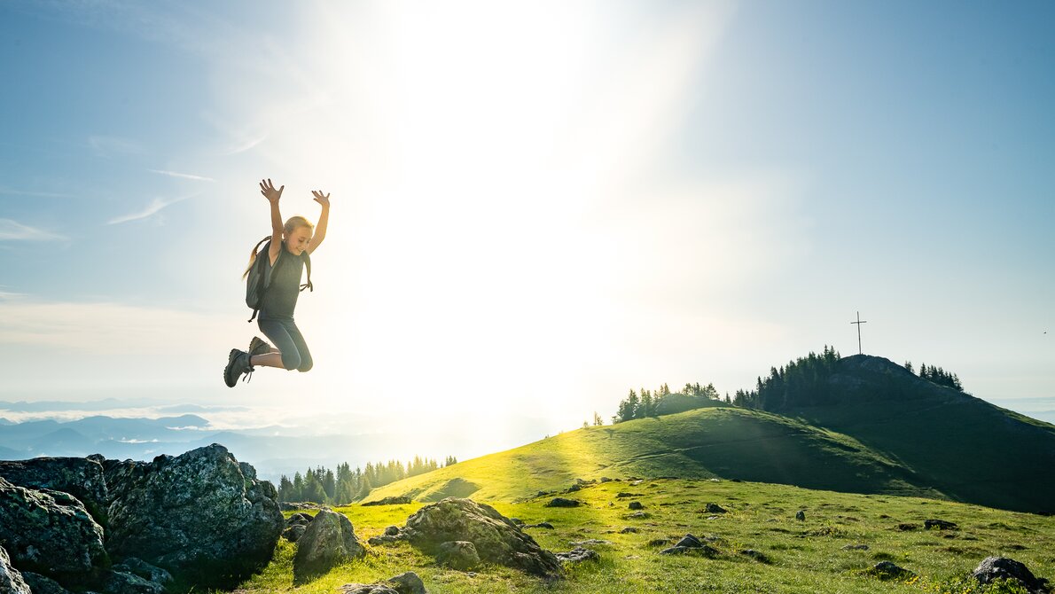 Ein Mädchen springt fröhlich in die Luft vor einer sonnigen Berglandschaft. | © Region Graz - Mias Photoart-43