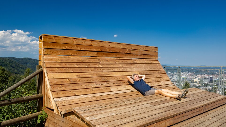 Ein Mann liegt entspannt auf einer hölzernen Liege mit Blick auf Graz. | © Graz Tourismus - Harry Schiffer