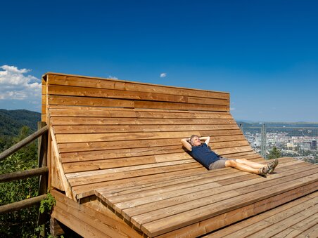Ein Mann liegt entspannt auf einer hölzernen Liege mit Blick auf Graz. | © Graz Tourismus - Harry Schiffer