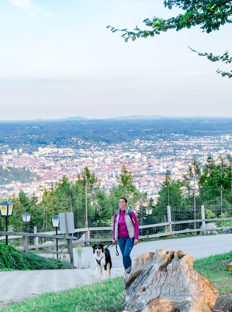 Eine Frau geht mit ihrem Hund auf einem Weg mit Aussicht über Graz. | © Graz Tourismus - Mias Photoart