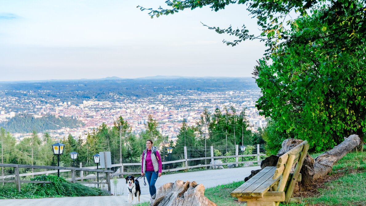 Eine Frau geht mit ihrem Hund auf einem Weg mit Aussicht über Graz. | © Graz Tourismus - Mias Photoart
