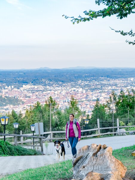 A woman walks with her dog on a path overlooking Graz. | © Graz Tourismus - Mias Photoart