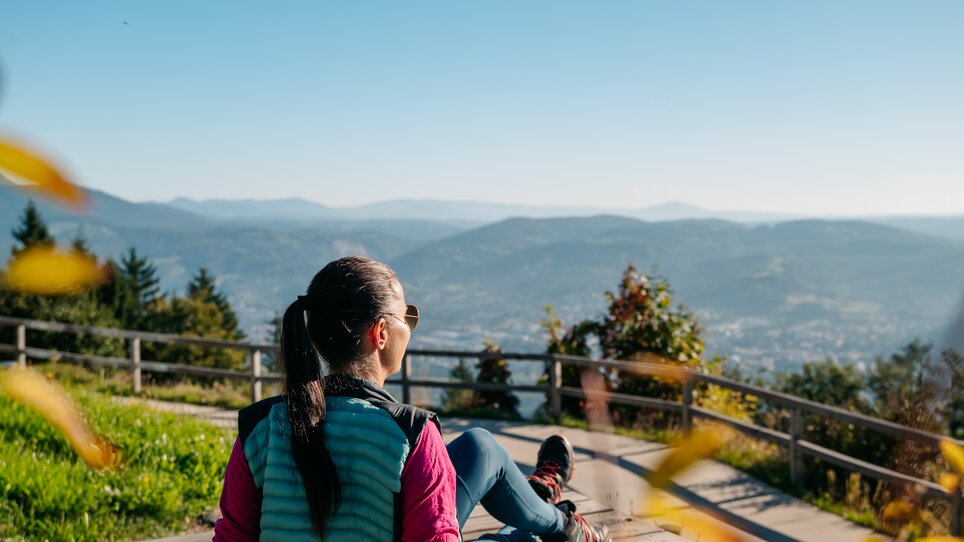 Frau sitzt auf einer Holzbank mit Blick über Graz und die Berge. | © Graz Tourismus - Mias Photoart