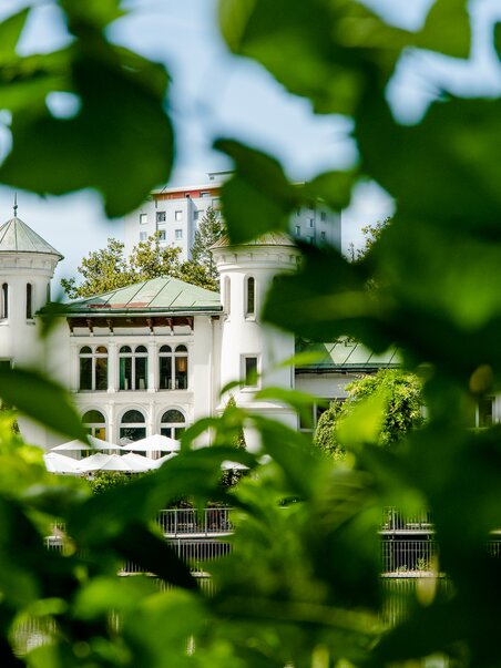 Un edificio sull’Hilmteich di Graz è incorniciato da foglie in primo piano. | © Graz Tourismus - Mias Photoart
