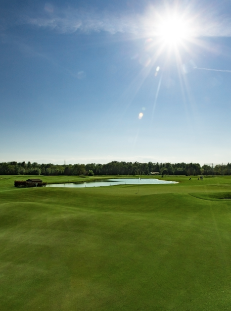 Ampio campo da golf con erba verde e specchio d'acqua sotto il sole splendente. | © GC Grazer MurAuen