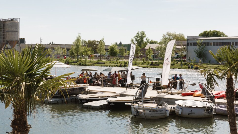 A busy jetty on the banks of the Mur in Graz with lots of people and boats. | © Die Flößerei