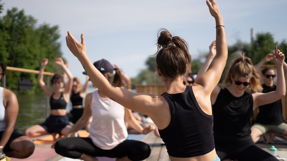 A group of people is taking part in a yoga class on a raft on the Mur River in Graz, offered by “Der Flößerei”. | © Robert Supnig