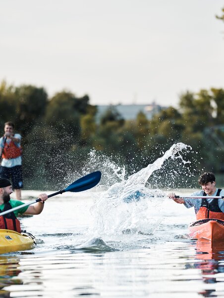 Zwei Kajakfahrer paddeln auf ruhigem Wasser, während ein Dritter auf einem SUP steht und zuschaut. | © Stadtstrand Graz - Stefan Leitner