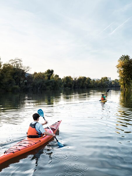 Zwei Personen im Kajak paddeln auf der ruhigen Mur umgeben von Bäumen. | © Stadtstrand Graz - Stefan Leitner