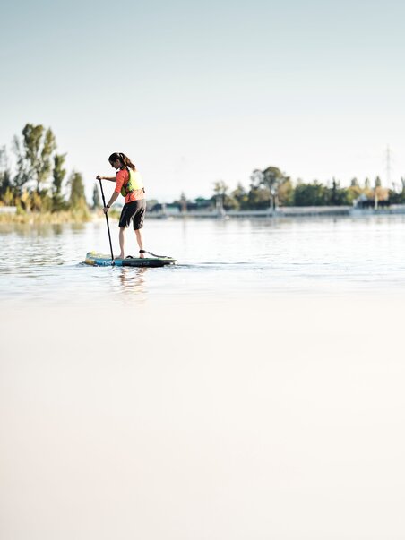 Ein Junge paddelt auf einem Stand-up-Paddleboard auf ruhig fließendem Wasser. | © Stadtstrand Graz - Stefan Leitner