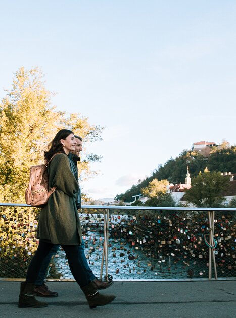 Ein Pärchen spaziert über eine Brücke in Graz mit Blick auf die Mur und den Grazer Uhrturm. | © Graz Tourismus