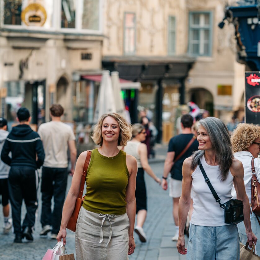 Zwei Frauen gehen fröhlich in Graz mit Einkaufstüten an einem belebten Tag spazieren. | © Graz Tourismus - Mias Photoart