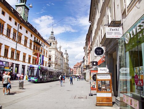 Blick auf eine belebte Straße in Graz mit Geschäften und Straßenbahn. | © sFachl