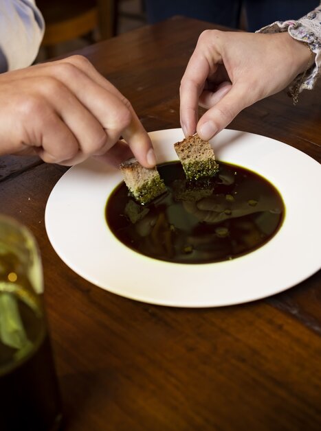 Due persone intingono il pane in un piatto bianco con olio di semi di zucca della Stiria. Accanto a loro c'è un piatto di semi di zucca e una bottiglia di vetro di olio di semi di zucca. | © Graz Tourismus - Werner Krug