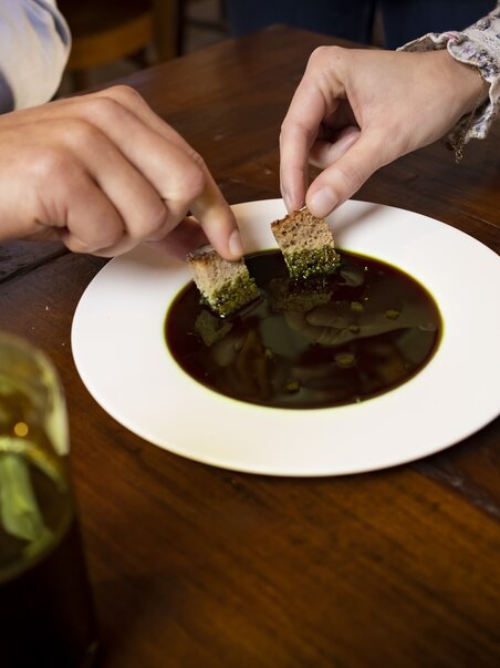 Zwei Personen tauchen Brot in einen weißen Teller mit steirischem Kernöl ein. Daneben steht ein Teller mit Kürbiskernen und eine Glasflasche mit Kernöl. | © Graz Tourismus - Werner Krug