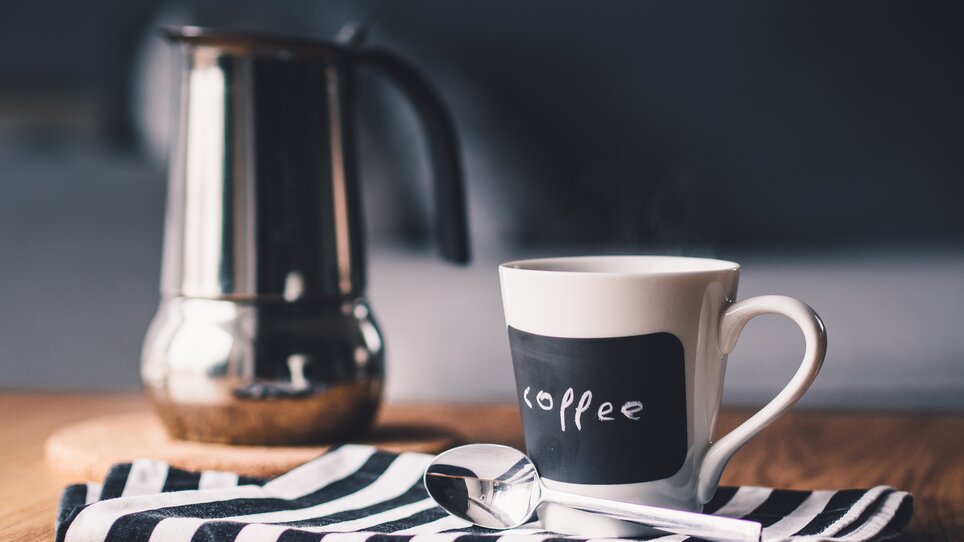 Eine Kaffeetasse steht neben einem Kaffeebereiter auf einem gestreiften Tuch. | © Pixabay