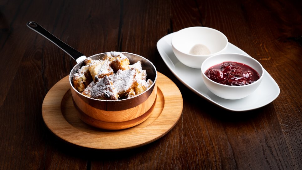 Kaiserschmarrn with sugar in a small pan, next to it is a bowl of stewed plums and an empty bowl.