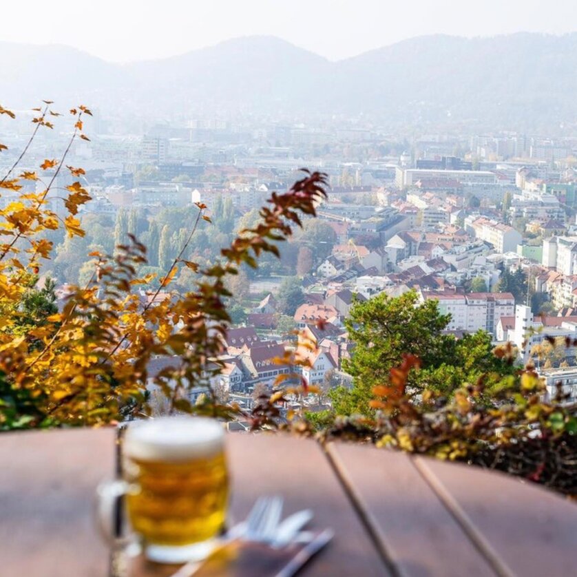 Restaurant SCHLOSSBERG I Biergarten I Essen & Trinken in Graz | © Restaurant Schlossberg Ein runder Tisch mit einem Glas Bier - im Hintergrund Aussicht auf die Stadt Graz. | © Restaurant Schlossberg