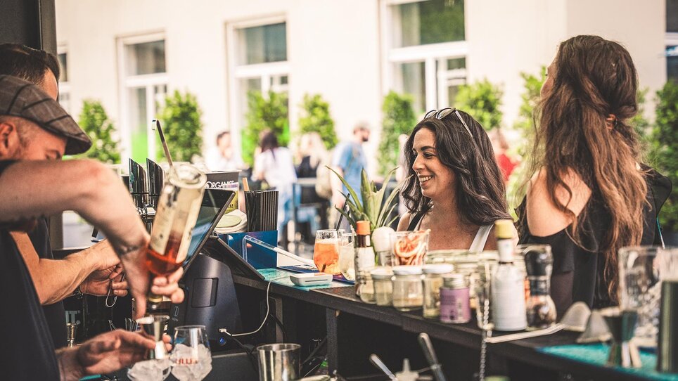 Zwei Frauen an einer Bar mit Getränken, ein Barkeeper bereitet ein Getränk vor. | © Viertel 4