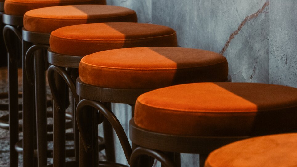 Reihe von Holzbarhockern mit orangefarbenen Sitzen an einer Wand. | © Ciccio