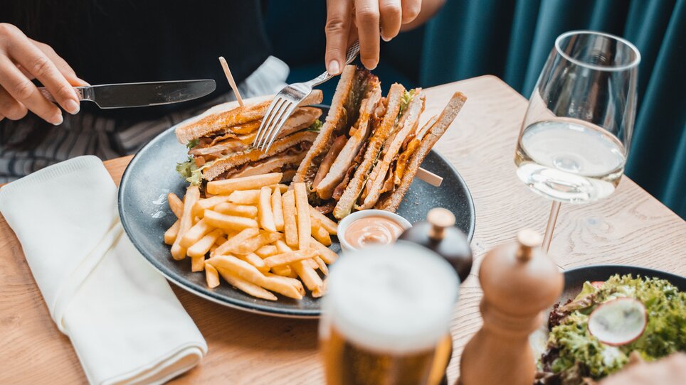 Eine Person im Restaurant schneidet ein Sandwich mit Pommes auf einem Teller, danben steht ein Glas Wein. | © Das Franz