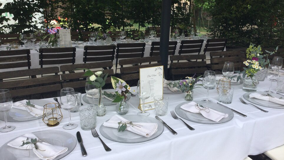 Tavolo elegantemente apparecchiato per evento all'aperto, adornato di fiori e menu. | © Das Lorenz