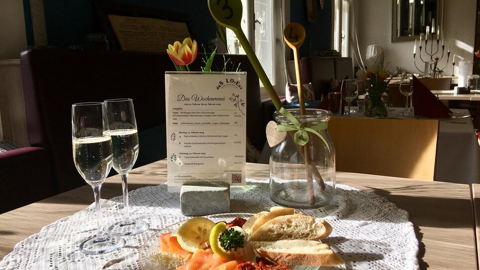 Un tavolo nel ristorante con un menù, vino frizzante e un piatto di cibo. | © Das Lorenz