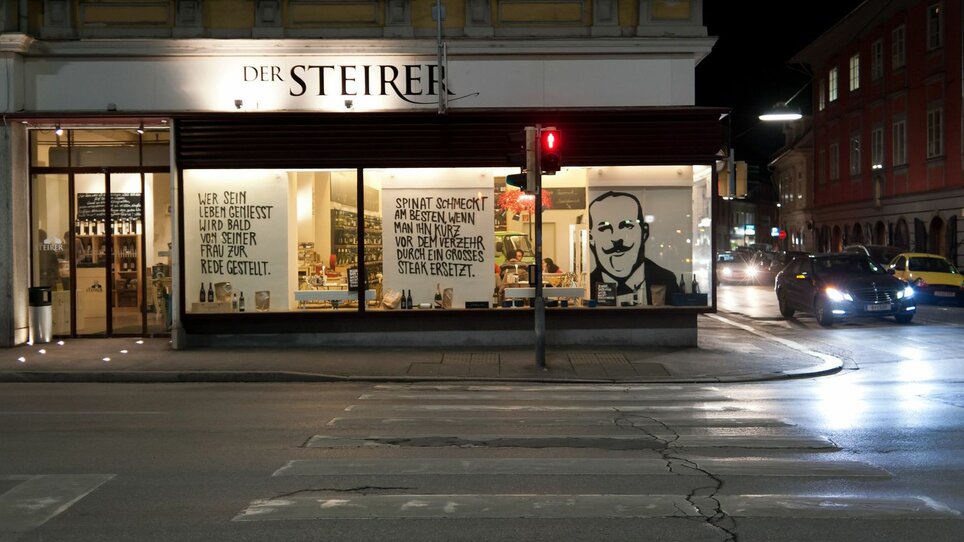Evening shot of the restaurant Der Steirer in Graz with inviting windows and a large mural. | © Weitzer