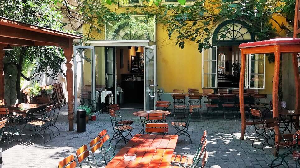 Gemütlicher Außenbereich mit Tischen und Stühlen, umgeben von Bäumen. | © Eschenlaube