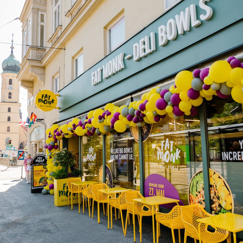 Außenansicht des Lokals FAT MONK Deli Bowls mit buntem Dekor und gelben Tischen und Stühlen im Außenbereich. | © Fat Monk – Deli Bowls