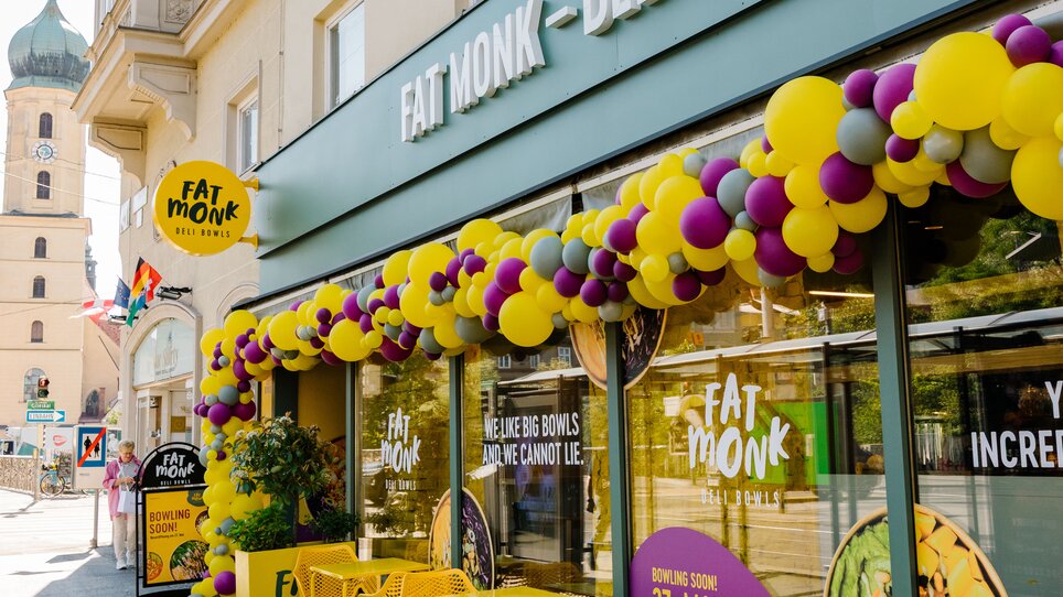 Außenansicht des Lokals FAT MONK Deli Bowls mit buntem Dekor und gelben Tischen und Stühlen im Außenbereich. | © Fat Monk – Deli Bowls