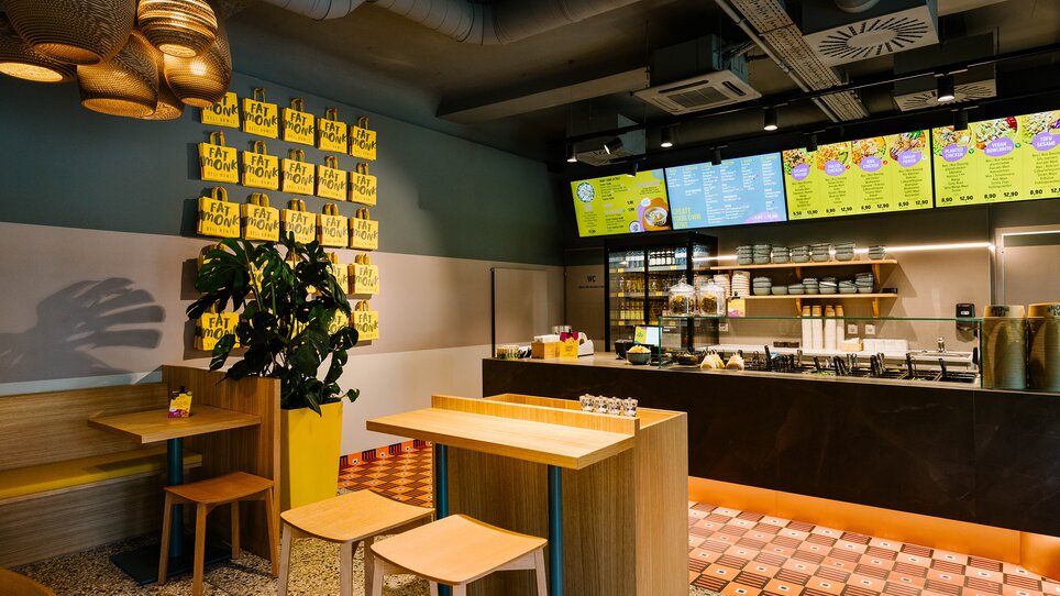 Innenansicht des Lokals Fat Monk Graz mit Holzmöbeln und farbenfrohen Menüs. | © Fat Monk – Deli Bowls