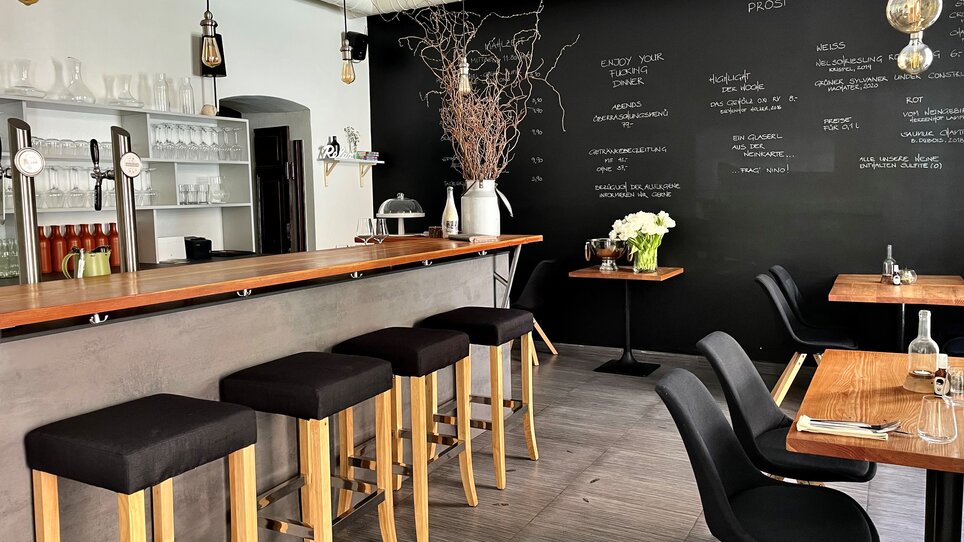 Ein modernes Restaurant mit einer Holzbar, Stühlen und einem schwarzen Wandbereich mit Speisen und Getränken. | © Gerüchteküche