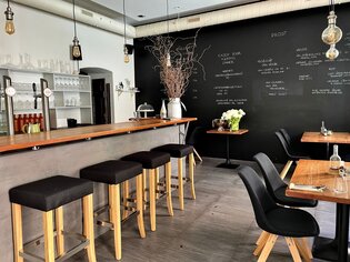 A modern restaurant featuring a wooden bar, stools, and a black wall with food and drink listings. | © Gerüchteküche